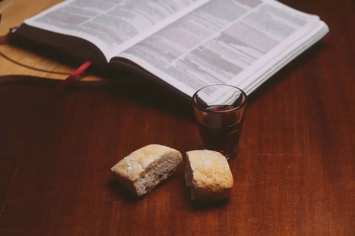 Bíblia aberta ao lado de pão e vinho sobre mesa de madeira, com vela acesa ao fundo