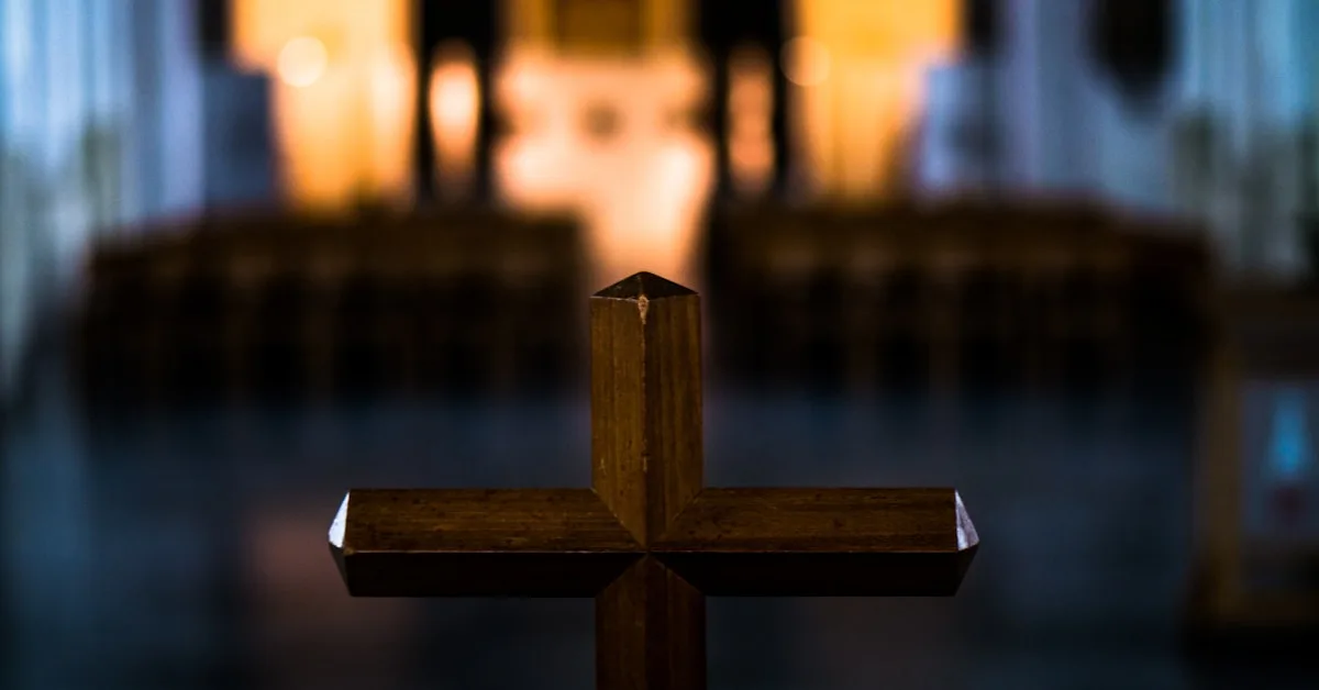 Uma cruz de madeira simples e rústica posicionada no interior de uma igreja iluminada de forma suave.
