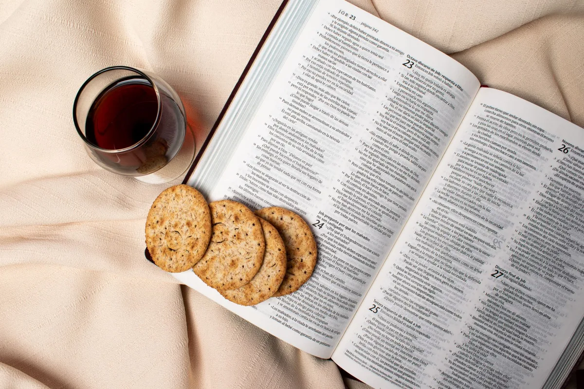 Bíblia aberta com vela acesa, pão e vinho sobre superfície escura, evocando os sacramentos
